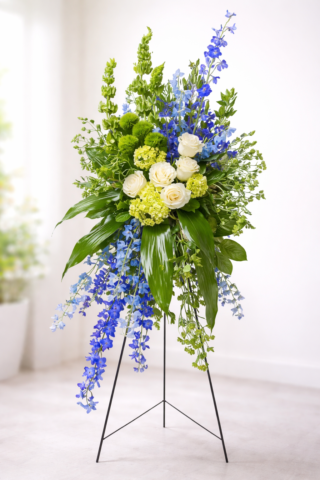 Graceful standing sympathy spray featuring white roses, green hydrangea, vibrant blue delphinium, textured green accents, and cascading greenery, arranged in an elegant vertical design on a green easel against a soft, neutral background.