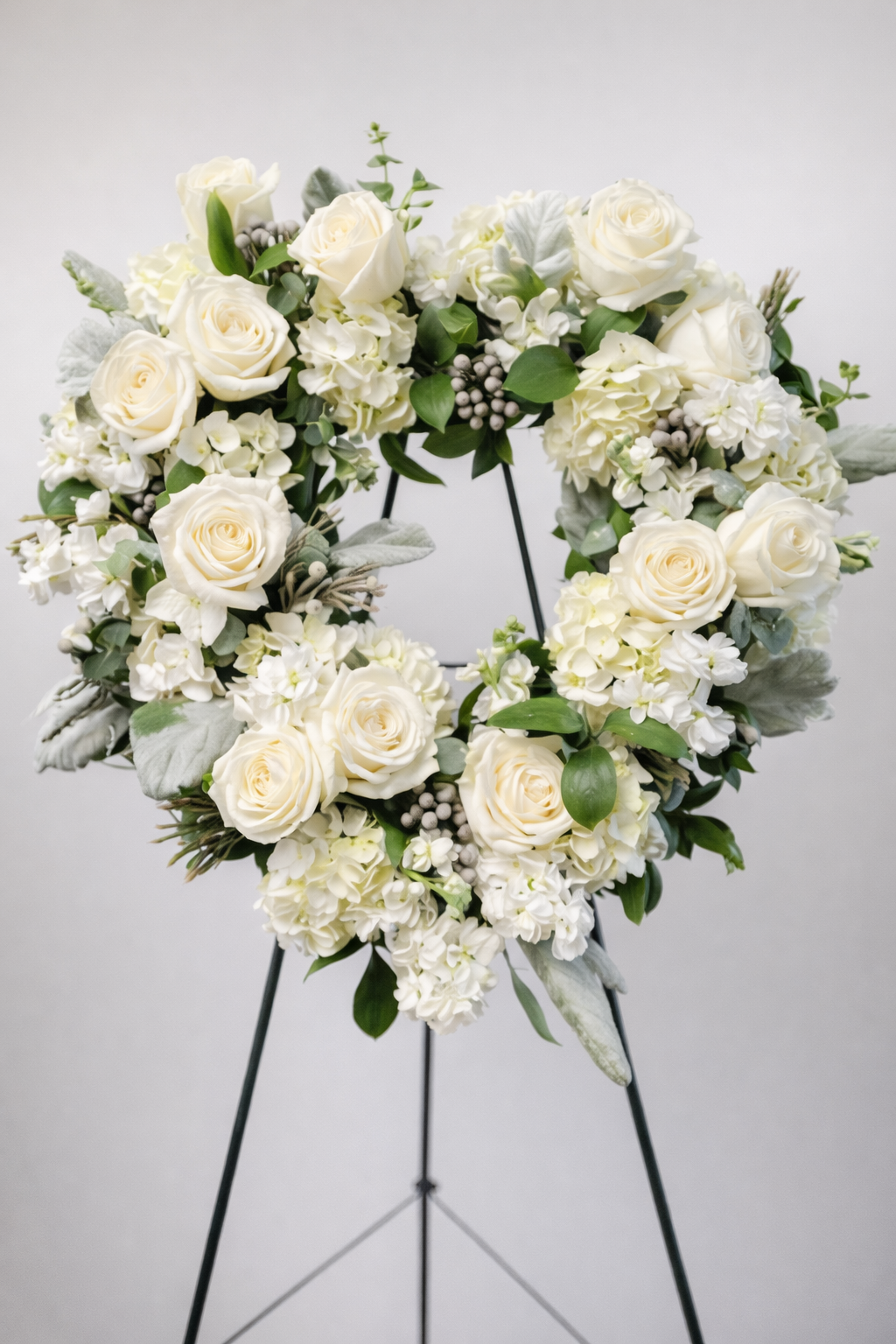 Elegant white sympathy open heart wreath featuring ivory roses, white hydrangea, soft textured greenery, and subtle berry accents, arranged in a classic heart design on a green easel against a neutral background.