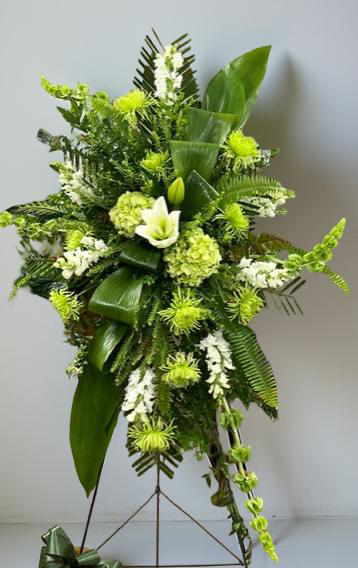Modern green standing sympathy spray featuring green hydrangea, textured green chrysanthemums, white snapdragons, and bold tropical foliage, arranged in a tall, asymmetrical design on a metal easel against a neutral background.