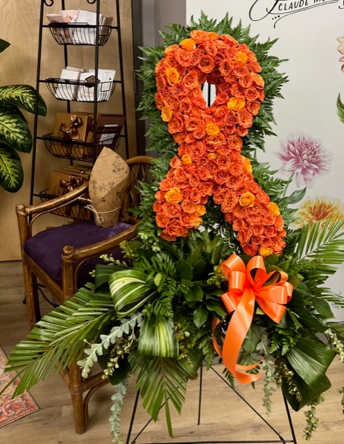 An orange rose awareness ribbon floral arrangement on a black metal stand, decorated with green foliage and a large orange bow, set in an indoor shop environment.