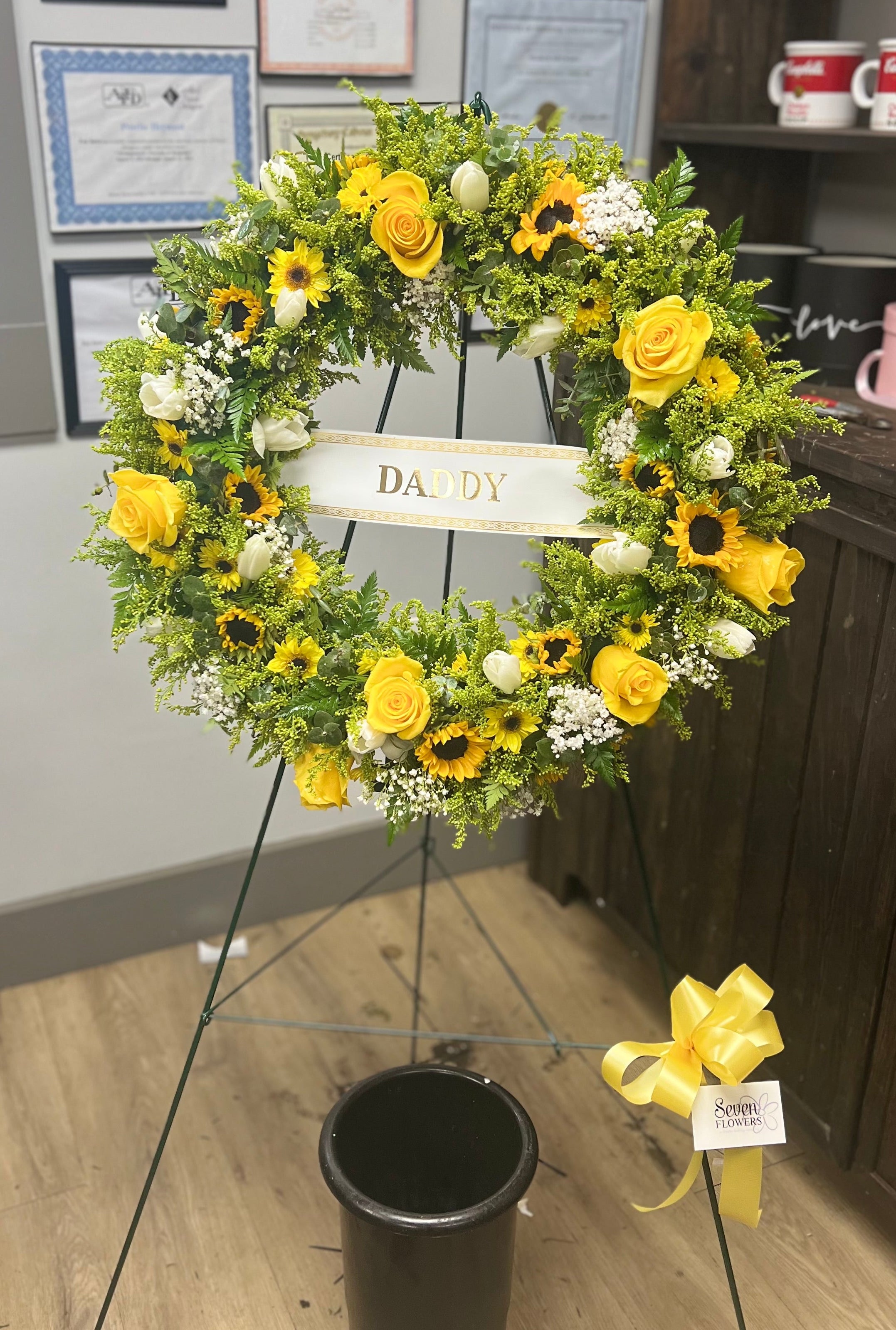 A yellow and sunflower memorial flower wreath on a stand, with a central sign reading &