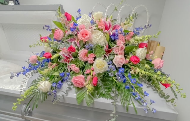 A casket spray featuring pink roses, blue delphinium, white hydrangea, and mixed greenery arranged in a full design across a gray casket, displayed against a gray background.