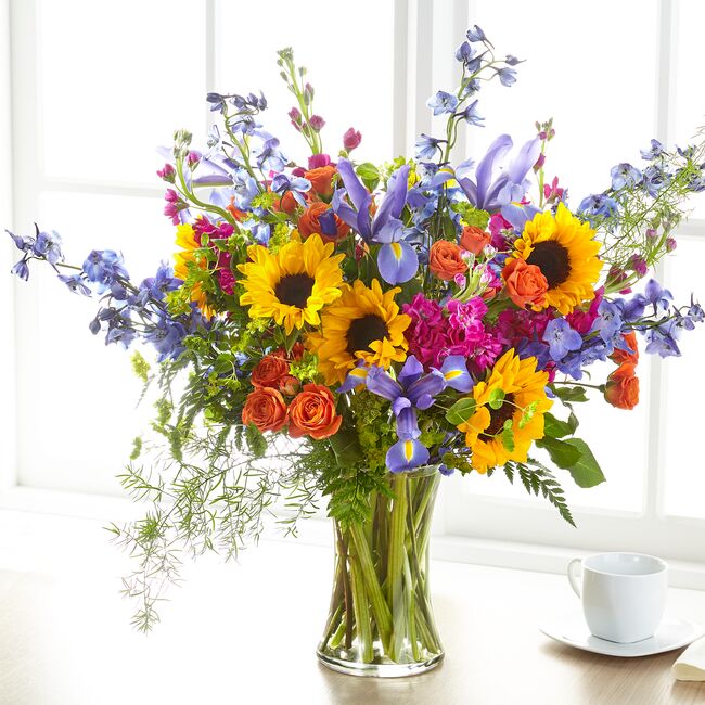 A vibrant bouquet of summer blooms including spray roses, delphiniums, irises, and sunflowers in a clear vase, placed on a table with a cup. Grandest size.