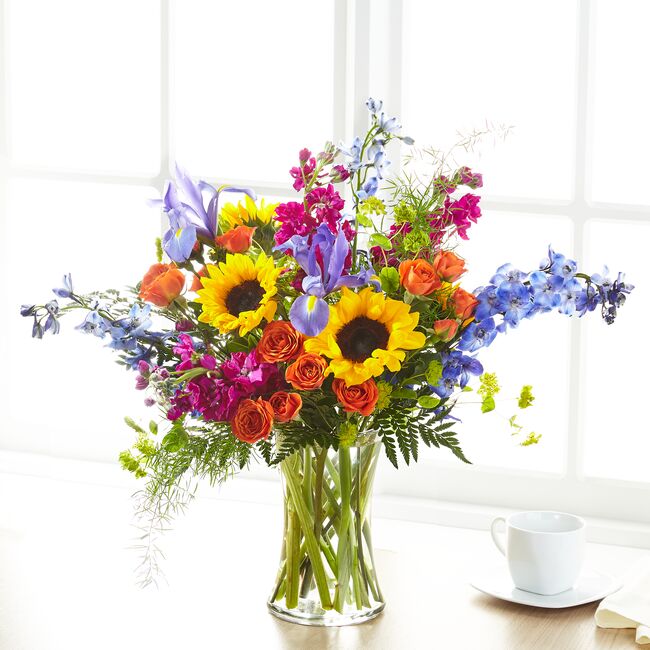 A vibrant bouquet of summer blooms including spray roses, delphiniums, irises, and sunflowers in a clear vase, placed on a table with a cup.