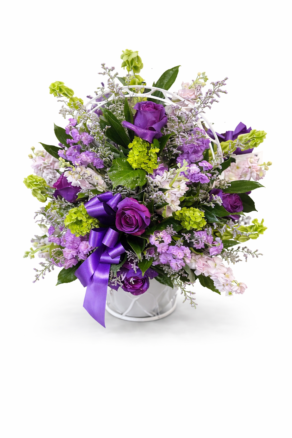 A sympathy flower basket featuring purple roses, lavender flowers, green hydrangea, and lush greenery arranged in a white handled basket with a purple ribbon, displayed against a clean white background.