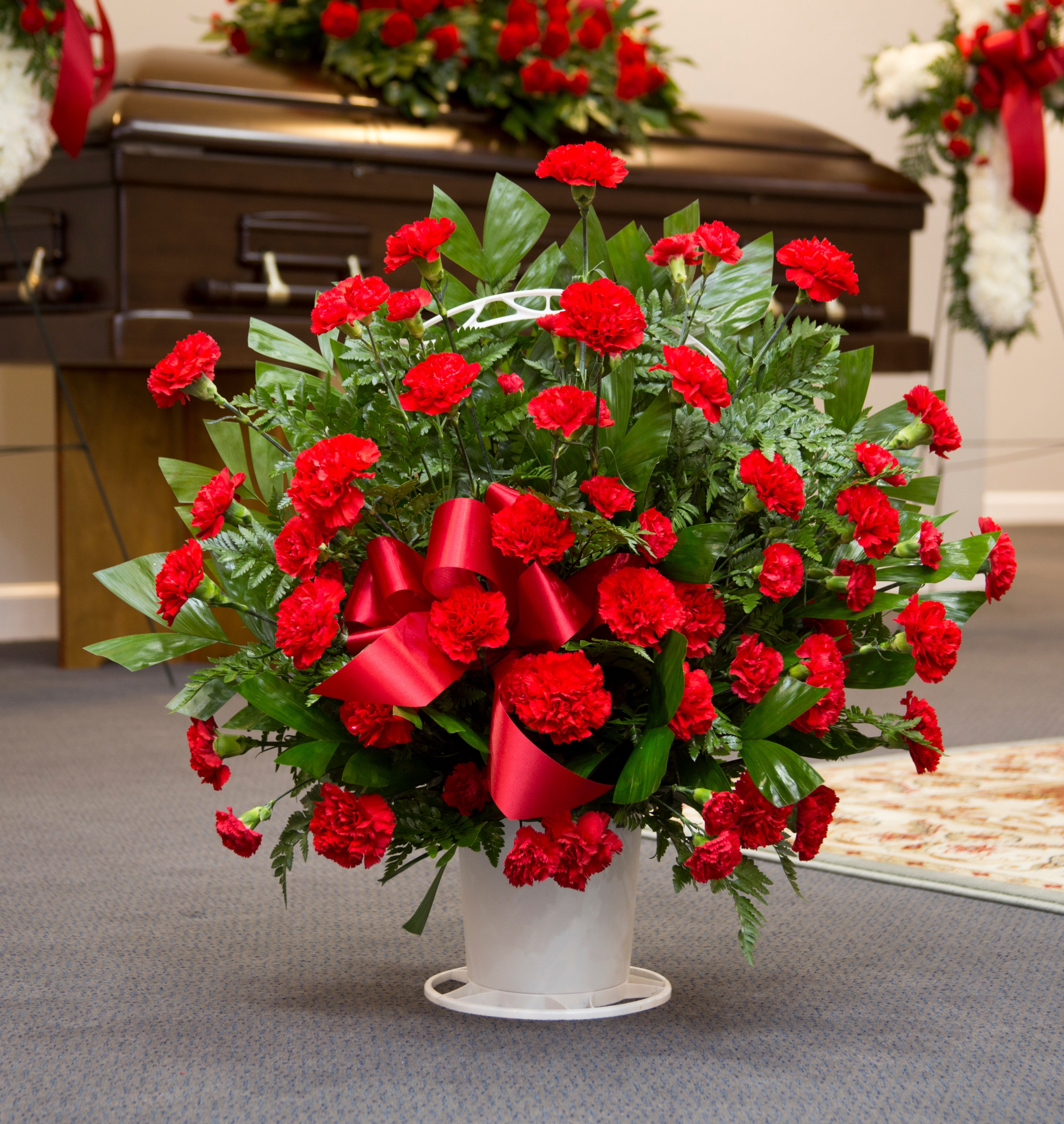 A large floral arrangement of red carnations with green foliage and a large red bow in a white pot, placed on a carpeted floor in front of a casket and white wreaths.