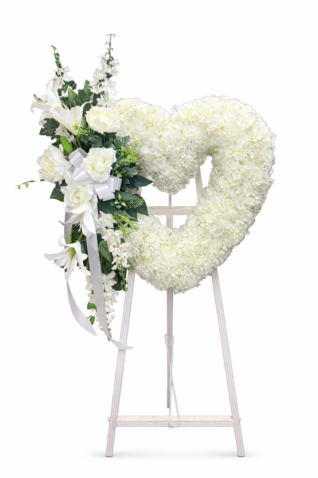 Elegant heart-shaped sympathy wreath made of artificial candlestick white carnations, accented with artificial white lilies, artificial white roses, artificial lush greenery, and a flowing white ribbon, displayed on a white easel against a clean neutral background.