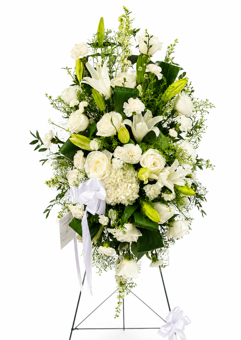 Classic white standing sympathy spray featuring white lilies, white roses, white cremon mums, carnations, delicate filler flowers, and lush palm and fern greenery, arranged in a full, cascading design on a green easel against a soft neutral background.