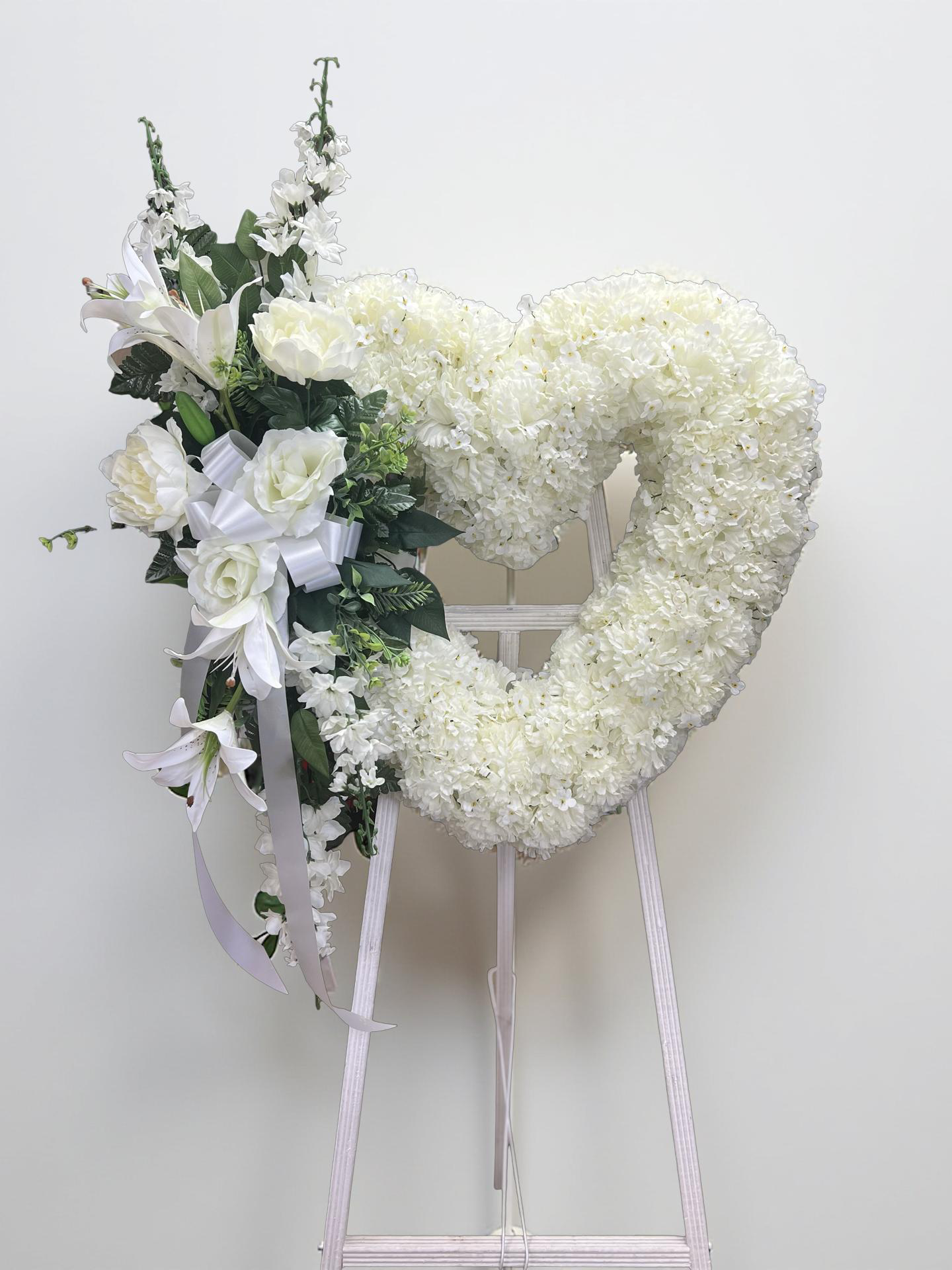 Elegant heart-shaped sympathy wreath made of artificial candlestick white carnations, accented with artificial white lilies, artificial white roses, artificial lush greenery, and a flowing white ribbon, displayed on a white easel against a clean neutral background.
