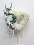 Elegant heart-shaped sympathy wreath made of artificial candlestick white carnations, accented with artificial white lilies, artificial white roses, artificial lush greenery, and a flowing white ribbon, displayed on a white easel against a clean neutral background.