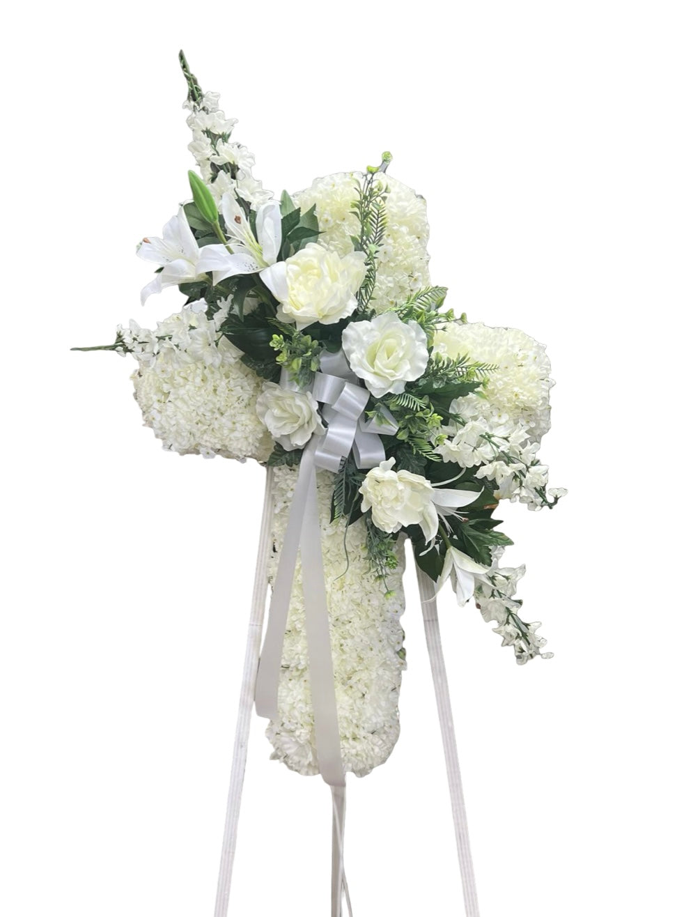 Elegant cross-shaped sympathy wreath made of artificial candlestick white carnations, accented with artificial white lilies, artificial white roses, artificial white delphiniums, artificial lush greenery, and a flowing white ribbon, displayed on a white easel against a clean neutral background.