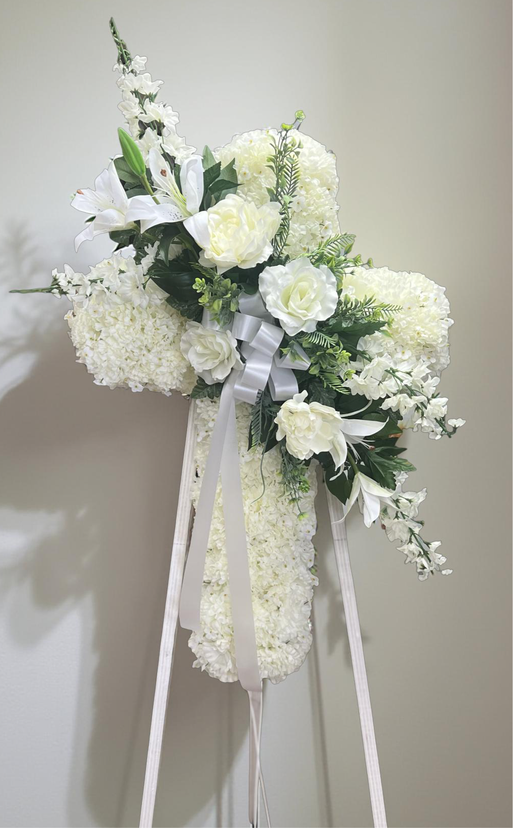 Elegant cross-shaped sympathy wreath made of artificial candlestick white carnations, accented with artificial white lilies, artificial white roses, artificial white delphiniums, artificial lush greenery, and a flowing white ribbon, displayed on a white easel against a clean neutral background.