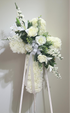 Elegant cross-shaped sympathy wreath made of artificial candlestick white carnations, accented with artificial white lilies, artificial white roses, artificial white delphiniums, artificial lush greenery, and a flowing white ribbon, displayed on a white easel against a clean neutral background.