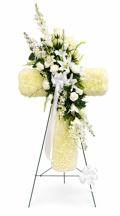Elegant heart-shaped sympathy wreath made of artificial candlestick white carnations, accented with artificial white lilies, artificial white roses, artificial lush greenery, and a flowing white ribbon, displayed on a white easel against a clean neutral background.