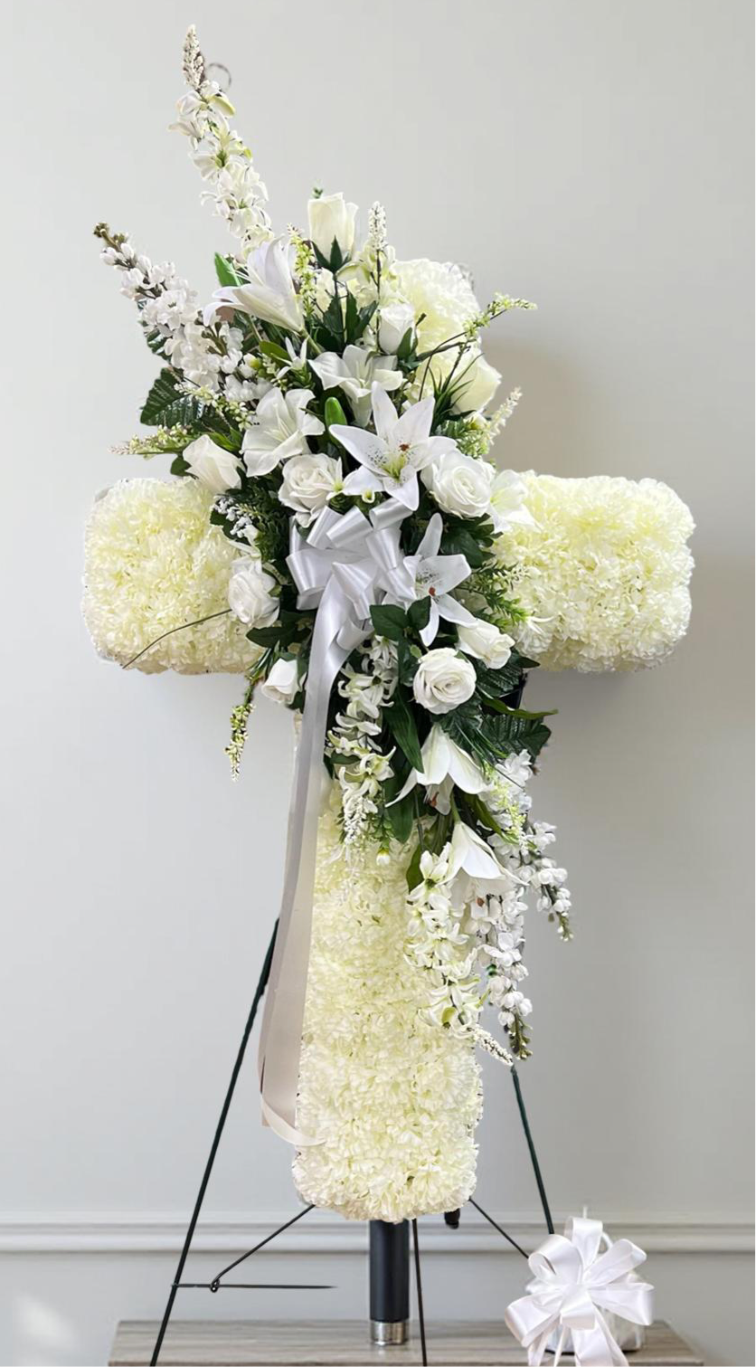 Elegant heart-shaped sympathy wreath made of artificial candlestick white carnations, accented with artificial white lilies, artificial white roses, artificial lush greenery, and a flowing white ribbon, displayed on a white easel against a clean neutral background.