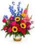 A colorful pedestal sympathy arrangement featuring bright yellow sunflowers, hot pink roses, blue delphinium, pink snapdragons, purple stock, lush greenery, and seasonal accent flowers arranged in an elegant white ceramic pedestal container with a gold base. The vibrant garden-style design symbolizes hope, joy, and a life beautifully remembered, making it suitable for funeral services, memorials, and celebrations of life.