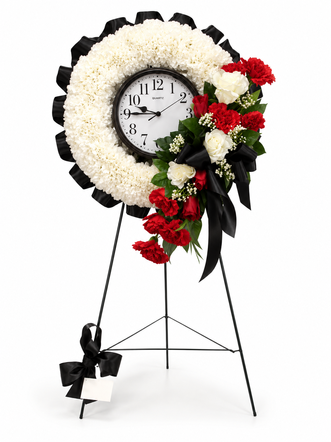 A memorial flower stand wreath with a round clock face in the center, surrounded by white carnations, red carnations, white roses, green leaves, and black ribbons on a black metal easel stand.