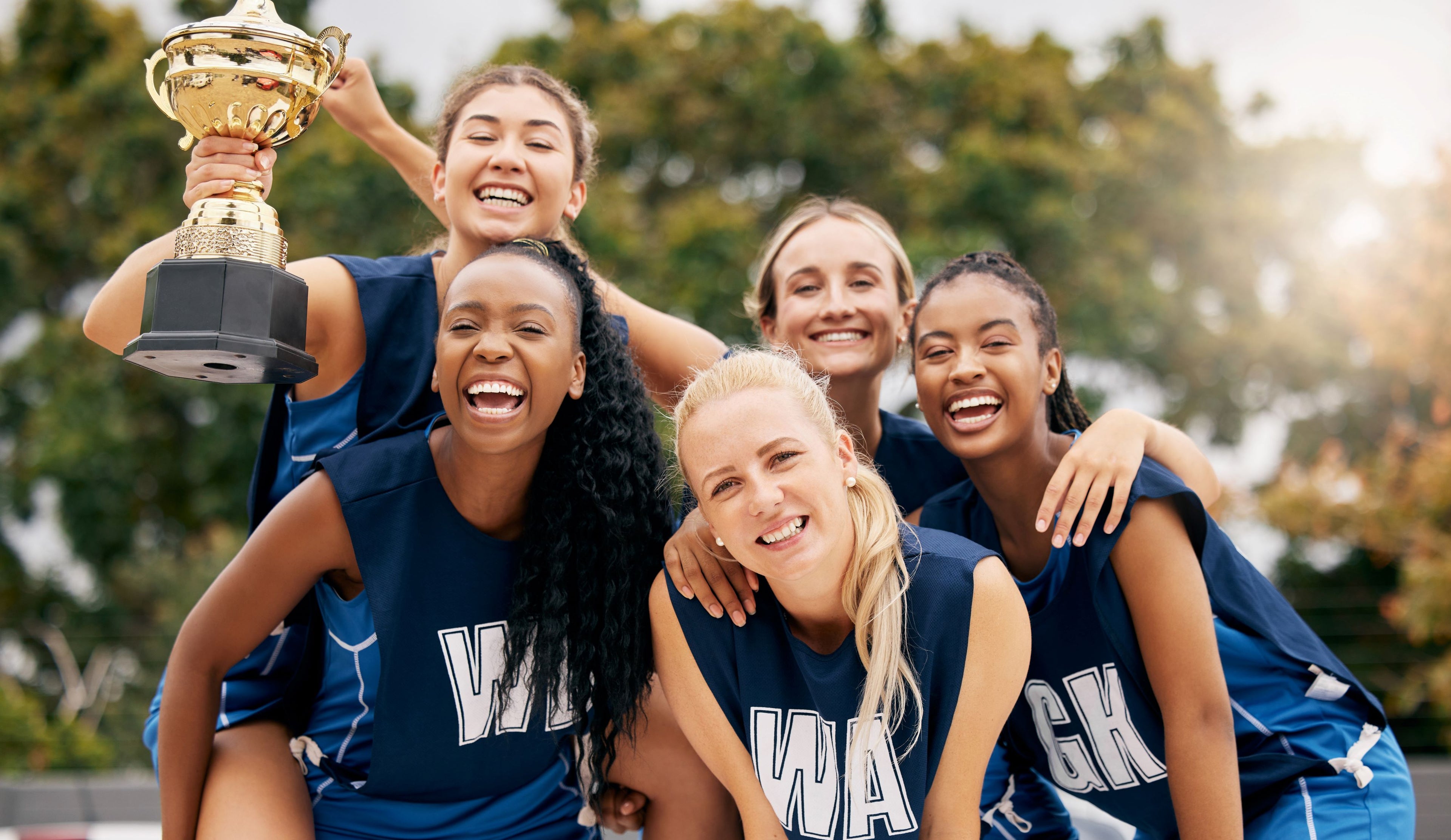 Group of smiling female athletes in matching blue uniforms celebrating together outdoors, one holding up a gold trophy while teammates gather closely, expressing joy and teamwork.