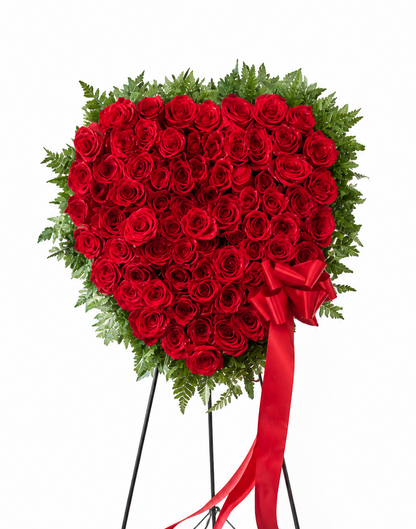 Heart-shaped standing sympathy wreath made entirely of deep red roses, framed with lush green foliage and accented with flowing red ribbon bows, displayed on a green easel against a white background.