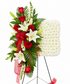 A floral arrangement on a black metal stand, featuring red roses, white lilies, green foliage, a red sheer ribbon, and a square white flower panel on the right, photographed on a plain white background.