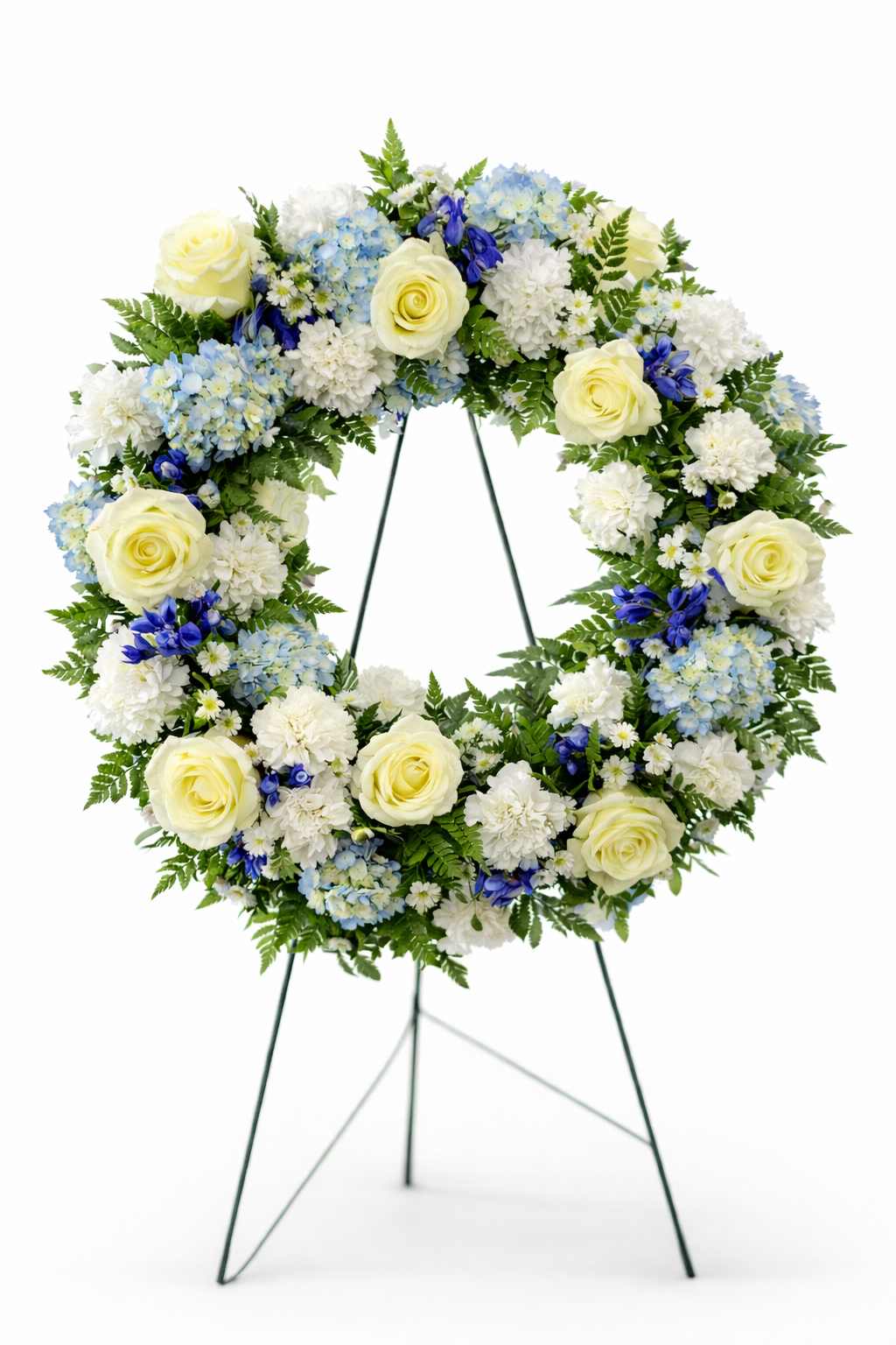 Classic sympathy wreath featuring ivory roses, white chrysanthemums, soft blue hydrangea, delicate blue delphiniums, and lush greenery, arranged in a full circular design on a green easel against a clean white background.