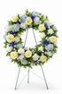 Classic sympathy wreath featuring ivory roses, white chrysanthemums, soft blue hydrangea, delicate blue delphiniums, and lush greenery, arranged in a full circular design on a green easel against a clean white background.