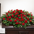 Traditional red carnation casket spray featuring 50 of deep red carnations accented with deep red mini carnations, lush green foliage, arranged in a full, symmetrical design atop a dark wood casket.