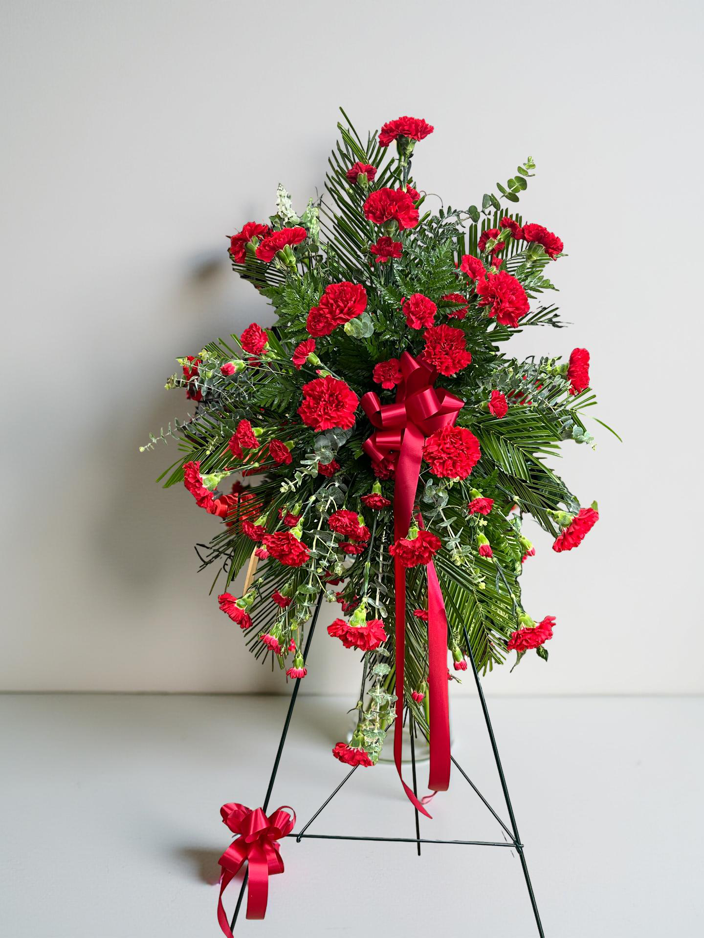 Standing sympathy spray featuring vibrant red carnations accented with lush evergreen foliage, finished with a flowing red satin ribbon, arranged in a classic vertical design on a green easel against a white background.