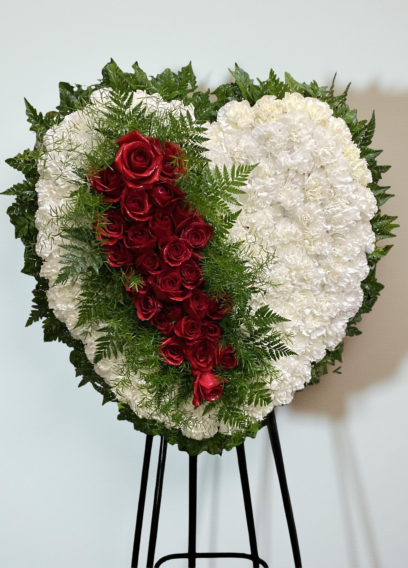 Heart-shaped sympathy wreath featuring white carnations accented with a cascading ribbon of deep red roses and lush greenery, arranged in a bold, elegant design on a green easel against a clean white background.