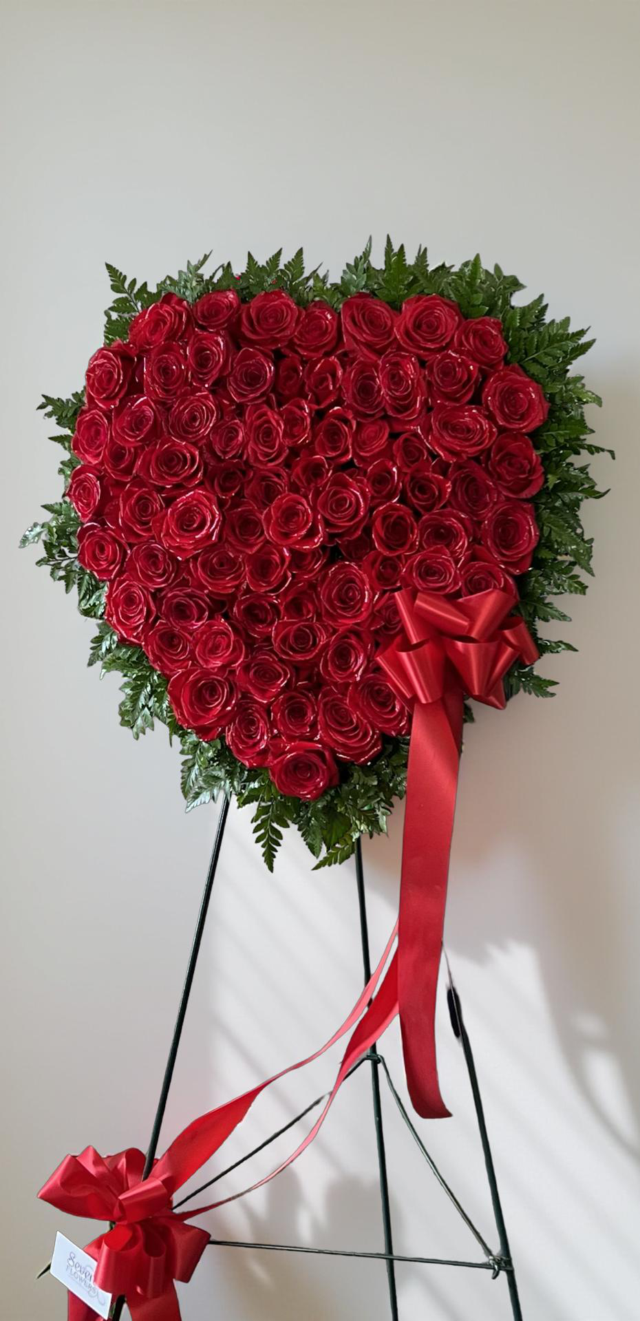 Heart-shaped standing sympathy wreath made entirely of deep red roses, framed with lush green foliage and accented with flowing red ribbon bows, displayed on a green easel against a white background.