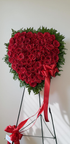 Heart-shaped standing sympathy wreath made entirely of deep red roses, framed with lush green foliage and accented with flowing red ribbon bows, displayed on a green easel against a white background.