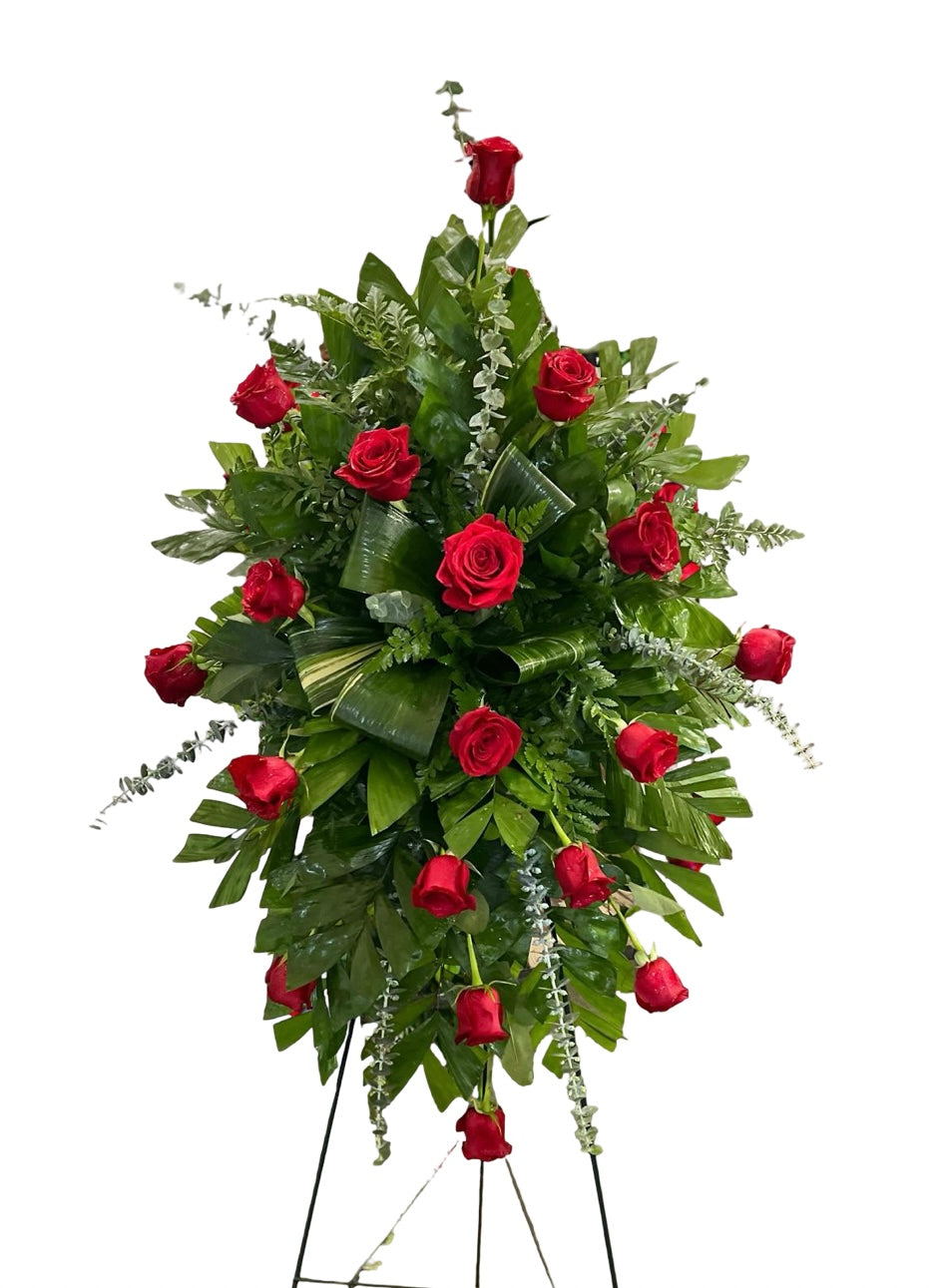 Elegant standing sympathy spray featuring deep red roses nestled among rich green foliage and cascading greenery, arranged in a classic vertical design on a green easel against a white background.