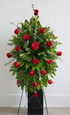 Elegant standing sympathy spray featuring deep red roses nestled among rich green foliage and cascading greenery, arranged in a classic vertical design on a green easel against a white background.
