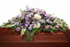 Soft pastel casket spray featuring lavender roses, purple and lavender carnations, white hydrangea, and flowing greenery, arranged in a full, graceful design atop a warm wood casket in a softly lit chapel setting.