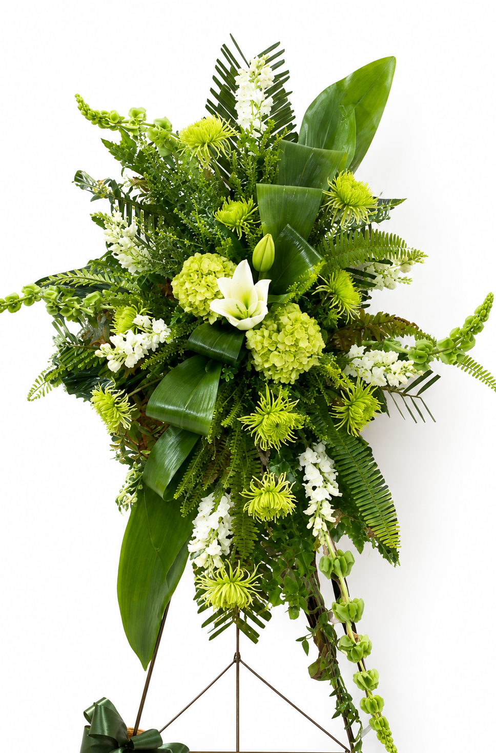 Modern green standing sympathy spray featuring green hydrangea, textured green chrysanthemums, white snapdragons, and bold tropical foliage, arranged in a tall, asymmetrical design on a metal easel against a neutral background.