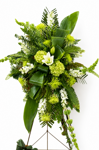 Modern green standing sympathy spray featuring green hydrangea, textured green chrysanthemums, white snapdragons, and bold tropical foliage, arranged in a tall, asymmetrical design on a metal easel against a neutral background.