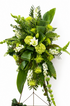 Modern green standing sympathy spray featuring green hydrangea, textured green chrysanthemums, white snapdragons, and bold tropical foliage, arranged in a tall, asymmetrical design on a metal easel against a neutral background.