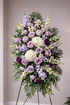 Elegant standing sympathy spray featuring lavender roses, white hydrangea, soft purple carnations and ivory roses, and lush palm and fern greenery, arranged in a full, layered design on a green easel against a neutral background.