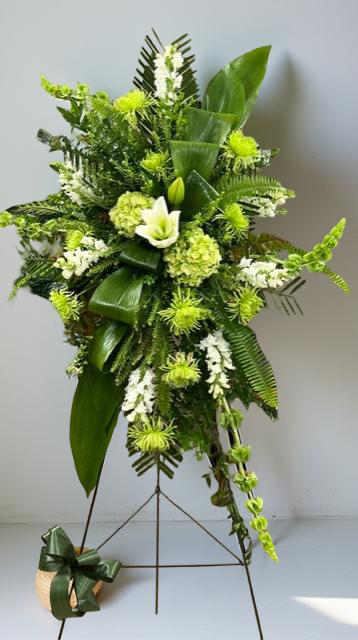 Modern green standing sympathy spray featuring green hydrangea, textured green chrysanthemums, white snapdragons, and bold tropical foliage, arranged in a tall, asymmetrical design on a metal easel against a neutral background.