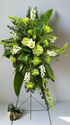 Modern green standing sympathy spray featuring green hydrangea, textured green chrysanthemums, white snapdragons, and bold tropical foliage, arranged in a tall, asymmetrical design on a metal easel against a neutral background.