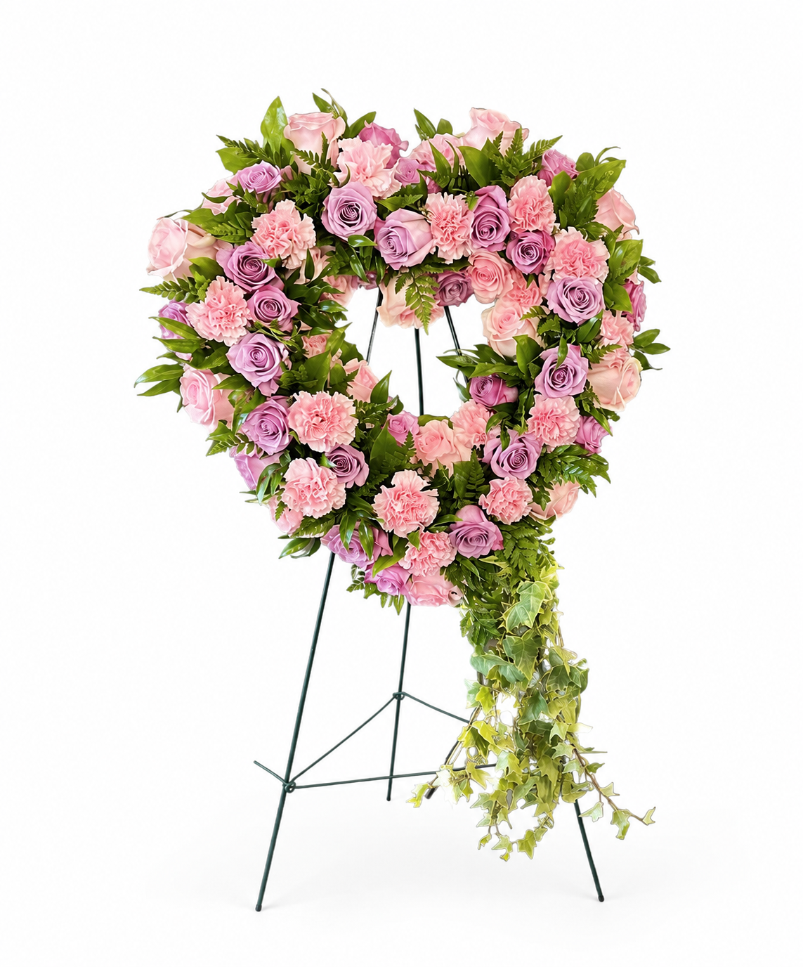 Heart-shaped sympathy wreath featuring soft pink and lavender roses, blush carnations, and lush green foliage, accented with cascading ivy, displayed on a green easel against a light neutral background.
