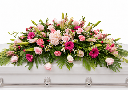 Graceful pink casket spray featuring soft pink roses, pink gerbera daisies, pale pink carnations, delicate filler flowers, and flowing greenery, arranged in a full, elegant design atop a white casket in a softly lit chapel setting.