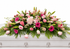 Graceful pink casket spray featuring soft pink roses, pink gerbera daisies, pale pink carnations, delicate filler flowers, and flowing greenery, arranged in a full, elegant design atop a white casket in a softly lit chapel setting.