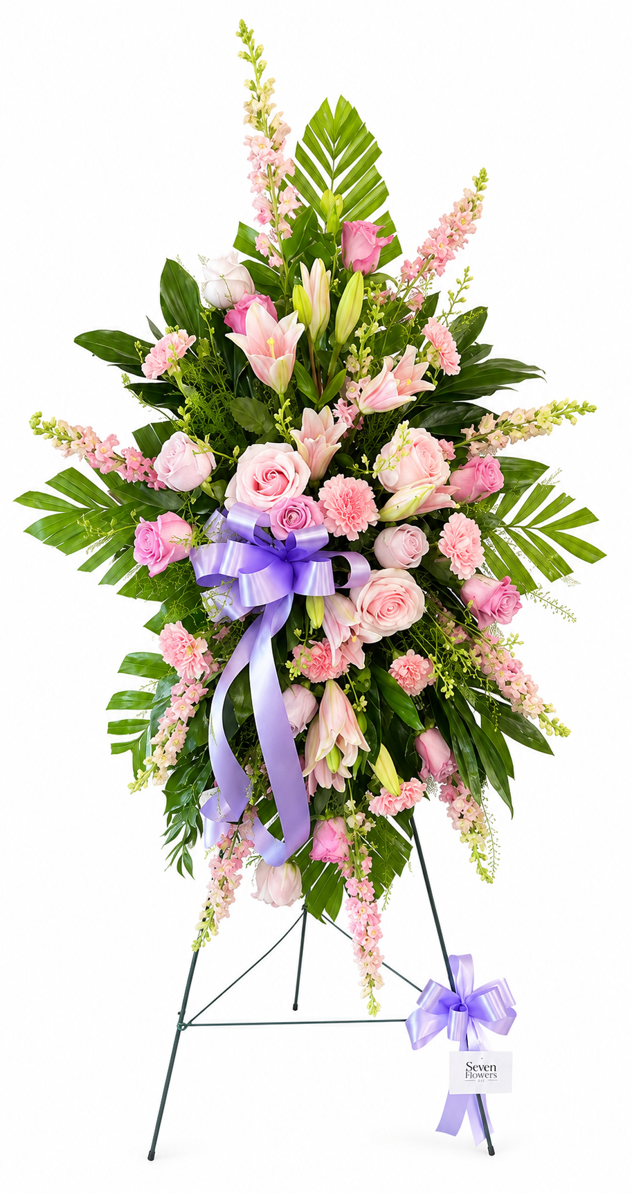 Soft pink standing sympathy spray featuring pink and lavender roses, blush lilies, pale carnations, and delicate filler flowers, accented with lush green foliage and finished with a lavender satin bow, arranged in a graceful vertical design on a green easel against a white background.