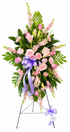 Soft pink standing sympathy spray featuring pink and lavender roses, blush lilies, pale carnations, and delicate filler flowers, accented with lush green foliage and finished with a lavender satin bow, arranged in a graceful vertical design on a green easel against a white background.