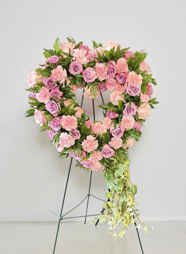 Heart-shaped sympathy wreath featuring soft pink and lavender roses, blush carnations, and lush green foliage, accented with cascading ivy, displayed on a green easel against a light neutral background.