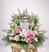 Graceful memorial urn arrangement featuring a silver urn surrounded by soft pink roses, pink gerbera daisies, white lilies, snapdragons, and lush greenery, designed in a full, elevated style on a wooden pedestal against a clean white background.