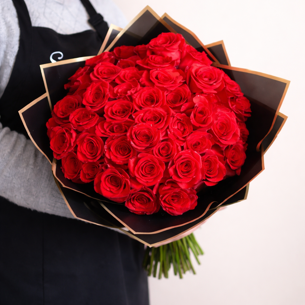 Bouquet of 50 red roses wrapped in black floral paper held by a person wearing an apron.