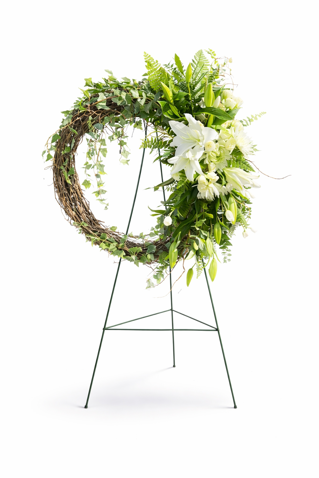 Natural sympathy wreath featuring a rustic grapevine base accented with white lilies, fresh green foliage, and trailing ivy, arranged in an asymmetrical design on a green easel against a clean white background.