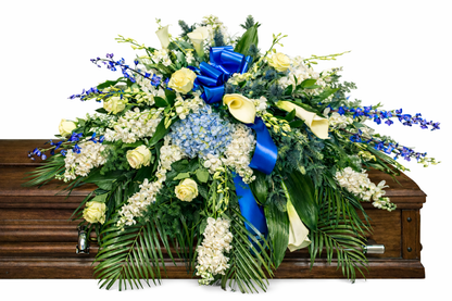 Elegant casket spray featuring white roses, white calla lilies, blue hydrangea, delicate blue delphiniums , and lush greenery, accented with a royal blue satin bow and arranged atop a wooden casket in a softly lit chapel setting.